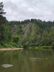 Fototapeta premium Serene River Landscape with Lush Greenery and Rocky Cliffs Under a Cloudy Sky, Perfect for Tourism