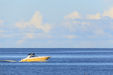 A sleek yellow motorboat with black canopy glides across tranquil deep blue ocean under clear sky with soft white clouds.