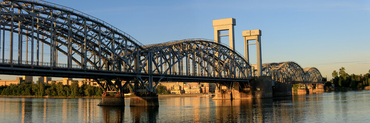 Wide panoramic view of Finland Railway Bridge in Saint Petersburg, with arched steel truss, lift towers, and golden reflections on Neva River.