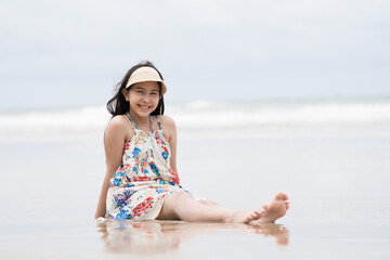 Happy smiling Asian girl sitting on sand and playing water on tropical beach in summer holiday with cloud, sky background