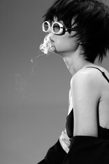 Black and white portrait of a young woman wearing round glasses, blowing smoke with a stylish edgy...