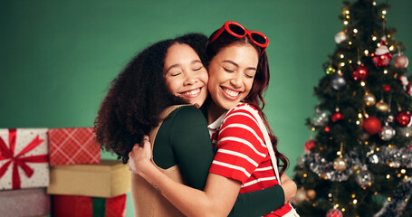 Women, hug and smile in Christmas studio with friends, holiday and festive cheer for bonding. Excited, love and embrace by tree for xmas celebration, presents or decoration on green background