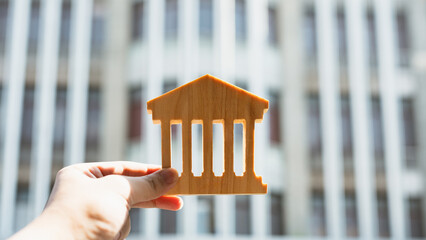 Wooden bank building model held by hand with city background, representing banking system,...