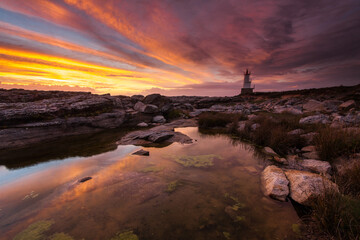 Lorient Morbihan Dramatic sky with red and orange tones at sunset, illuminating a lighthouse on rocky coast.