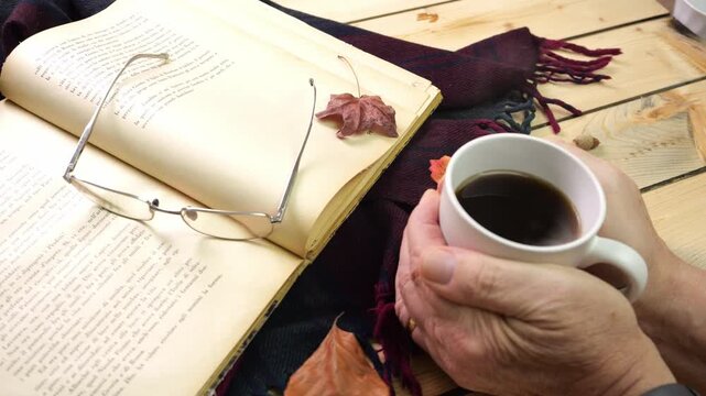 Uomo si scalda le mani sulla tazza di te caldo fumante, libro e occhiali con foglie secche, autunno