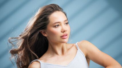 Young woman with healthy skin and flowing hair poses gracefully against a soft blue backdrop,...