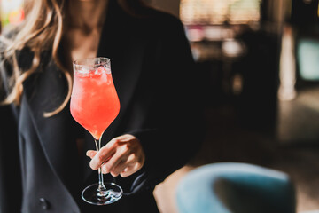 Hand holding a stemmed glass with red cocktail and ice, black outfit visible, blurred indoor background suggests bar or restaurant setting