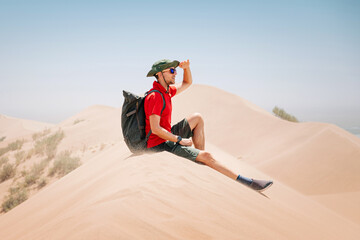 Tourist with backpack and hat sitting on sand dune shielding eyes from sun while exploring desert landscape