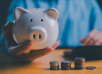 Close up of hand holding piggy bank with stacked coins on wooden table, symbolizing savings, financial planning, investment growth, money management, and smart economic budgeting