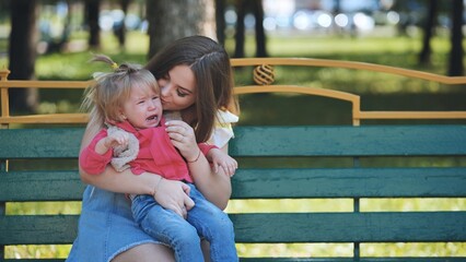 Young mother calming down her crying toddler daughter while sitting on a bench in a park during a...