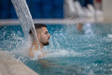 Man standing under a hydrotherapy waterfall in a spa pool, receiving water massage on his head and...