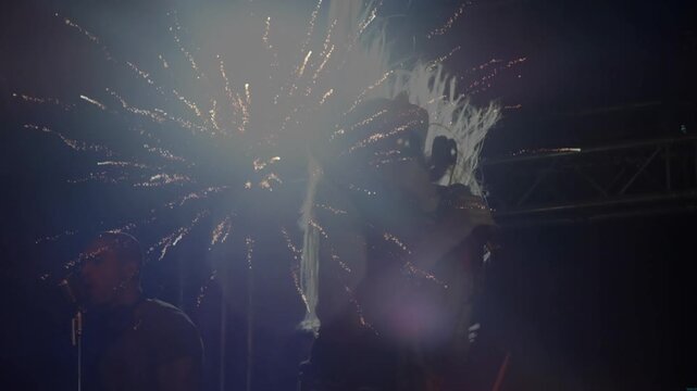 Fireworks igniting behind female singer on stage singing, flipping hair, unveiling band silhouette
