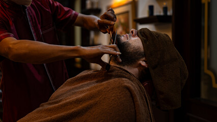 A skilled barber carefully trims a client’s beard with scissors in a barbershop. The man relaxes under a towel during precise grooming, showcasing traditional male care, style, and professional crafts