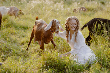 Smiling woman with dairy goats on organic pasture at sunset countryside
