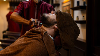 A barber trims a client’s beard with scissors in a barbershop. The man relaxes under a towel during the grooming session, highlighting precision, traditional craftsmanship, and professional male care.