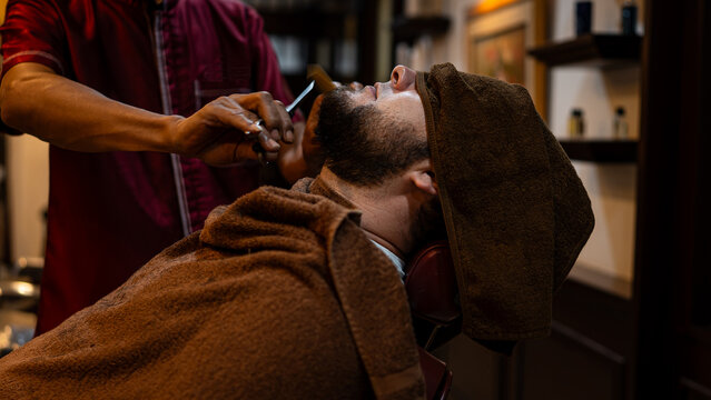 A barber carefully trims a client’s beard with scissors and a comb in a barbershop. The man relaxes under a towel during grooming, showcasing traditional craftsmanship, precision, and professional mal