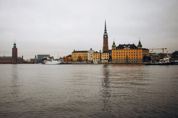 Naklejka premium Stockholm's Riddarholmen Church and historic buildings on the waterfront, with a boat on the water and the city hall in the distance.