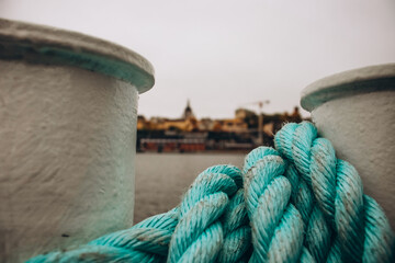 A close-up of a vibrant blue rope wrapped around a mooring bollard, with a blurred view of Stockholm's city skyline and water in the background.
