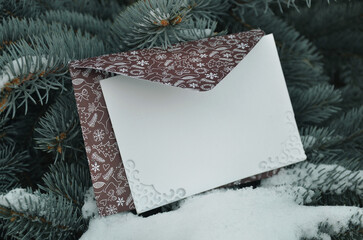 Brown envelope and white blank card against the background of snowcovered blue spruce christmas...