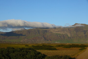 Wide Icelandic landscape with dramatic mountain ridges, low clouds stretching across the peaks, and a dirt road leading through green and brown fields in the evening light
