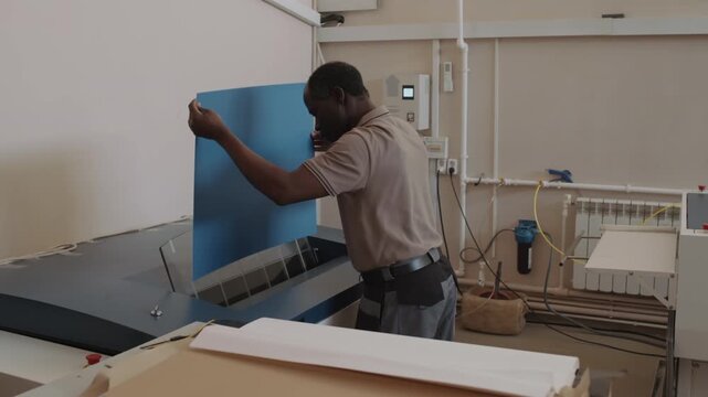 Senior African American man working in modern printing house putting sheet of blue paper into printer