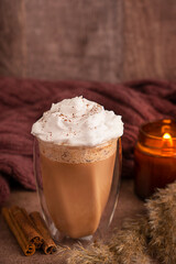Cozy autumn spiced latte coffee decorated with whipped cream topping served in glass on brown table with cinnamon sticks, dried plants and glowing candle as hot drink for comfortable breakfast