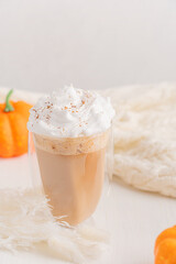 Hot pumpkin spice latte or coffee drink decorated with whipped cream topping served in drinking glass on white wooden table with cozy knitted blanket for comfortable aromatic autumn breakfast