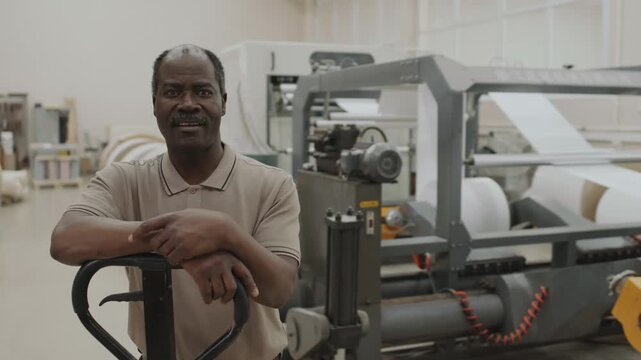 Medium portrait of senior African American man working in print shop standing against paper cutting machine smiling at camera