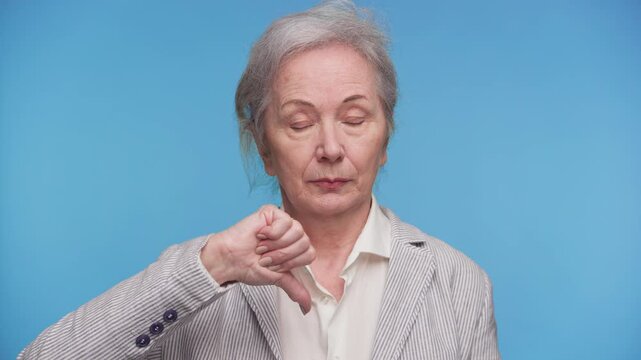 Serious senior woman giving thumbs down gesture on blue
