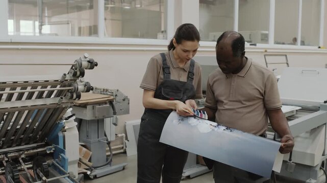 Medium long shot of mature Black man and young Caucasian woman working in print shop checking colors of printout