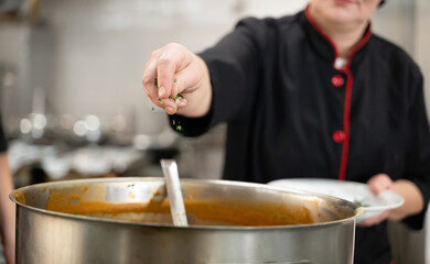 Close up of a chef in black uniform adding fresh herbs to a large pot of soup in a professional...