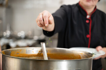 Close up of a chef in black uniform adding fresh herbs to a large pot of soup in a professional...