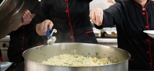 Group of chefs in black uniforms preparing rice in a large pot in a commercial kitchen. Female...