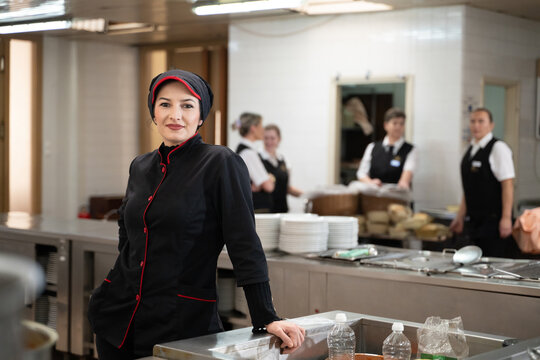 Portrait of a female chef in a black uniform with red details smiling in a professional restaurant kitchen. Image highlights hospitality staff, confidence, and culinary professionalism.