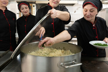 Group of chefs in black uniforms preparing rice in a large pot in a commercial kitchen. Female...