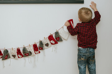 Little boy in red plaid shirt hanging Christmas gift bags on string garland from behind