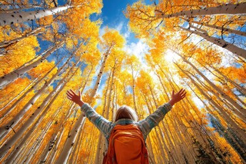 Adventurous child raising arms amidst tall golden yellow autumn trees under vibrant blue sky, exploring nature and celebrating fall foliage in a forest