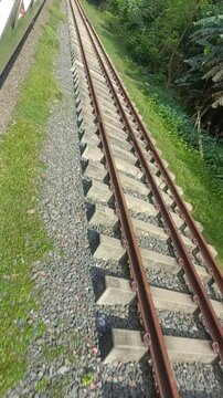 "A close-up perspective of parallel railway tracks captured from a moving train. The image shows steel rails, concrete sleepers, gravel, and green surroundings, representing travel, transportation