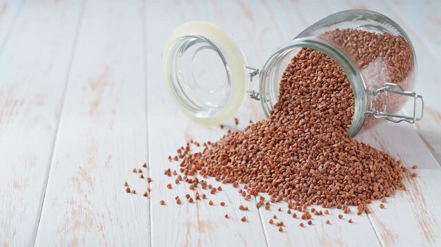 buckwheat spill out of a glass storage jar a light kitchen table. Raw buckwheat are scattered out of glass jar.
