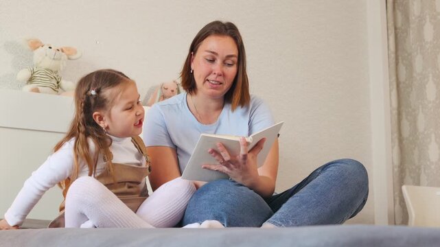 A caregiver helps a small girl read her first book in a bright room. Concept of preschool learning and child development. Bottom view.