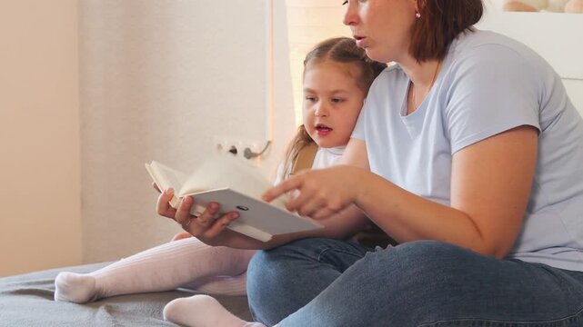A preschool girl and her mother read a fairytale together on a bed. Early reading skills and home-based development. Close up.