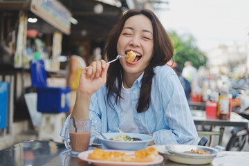 Smiling Asian woman eating Thai breakfast at local street food market, authentic lifestyle and happiness concept.