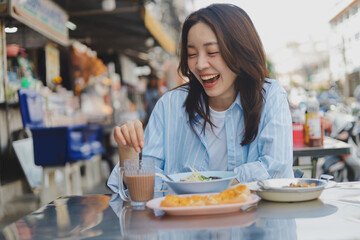 Smiling Asian woman eating Thai breakfast at local street food market, authentic lifestyle and happiness concept.
