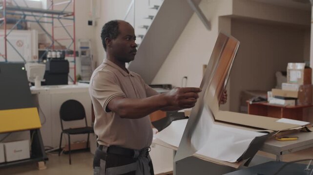 Senior African American male print shop worker taking sheet of paper then putting it into UV printer
