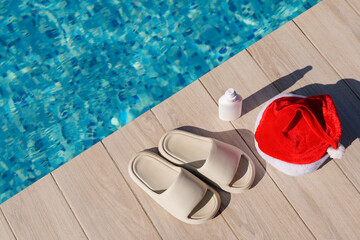 A high-angle shot of a swimming pool edge with a Christmas hat, a white lotion bottle, and a pair of light-colored slippers on a wooden deck. The image has a sunny, relaxed holiday feel.