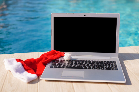 A laptop with a blank screen sitting next to a red Christmas hat on a wooden deck by a swimming pool.