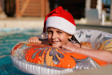 A happy young girl wearing a Christmas hat smiles while floating on an inflatable ring in a...