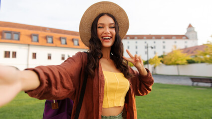 Positive young lady wear straw hat and smiling showing v-sign taking selfie outdoors, walking in...