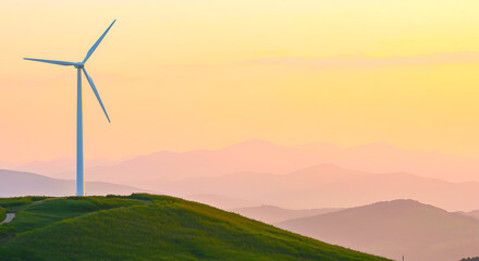 Obraz premium Majestic wind turbine stands tall on a grassy hilltop against a vibrant orange and yellow sunset sky with layered mountains