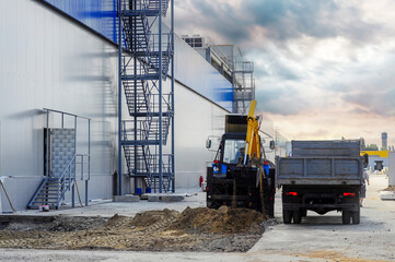 Backhoe and dump truck working on a construction site next to a large industrial building.